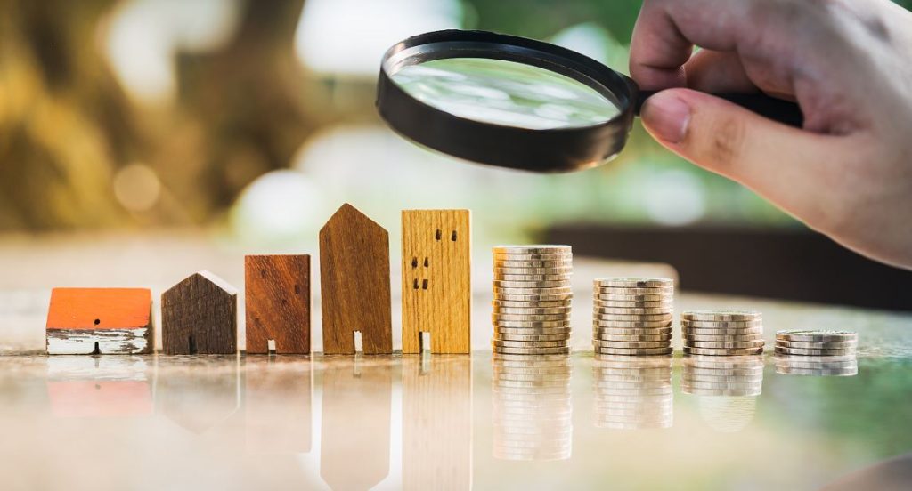 Main Tenant Une Loupe Qui Regarde Des Petites Maisons Et De L'argent, Illustre L'investissement Immobilier