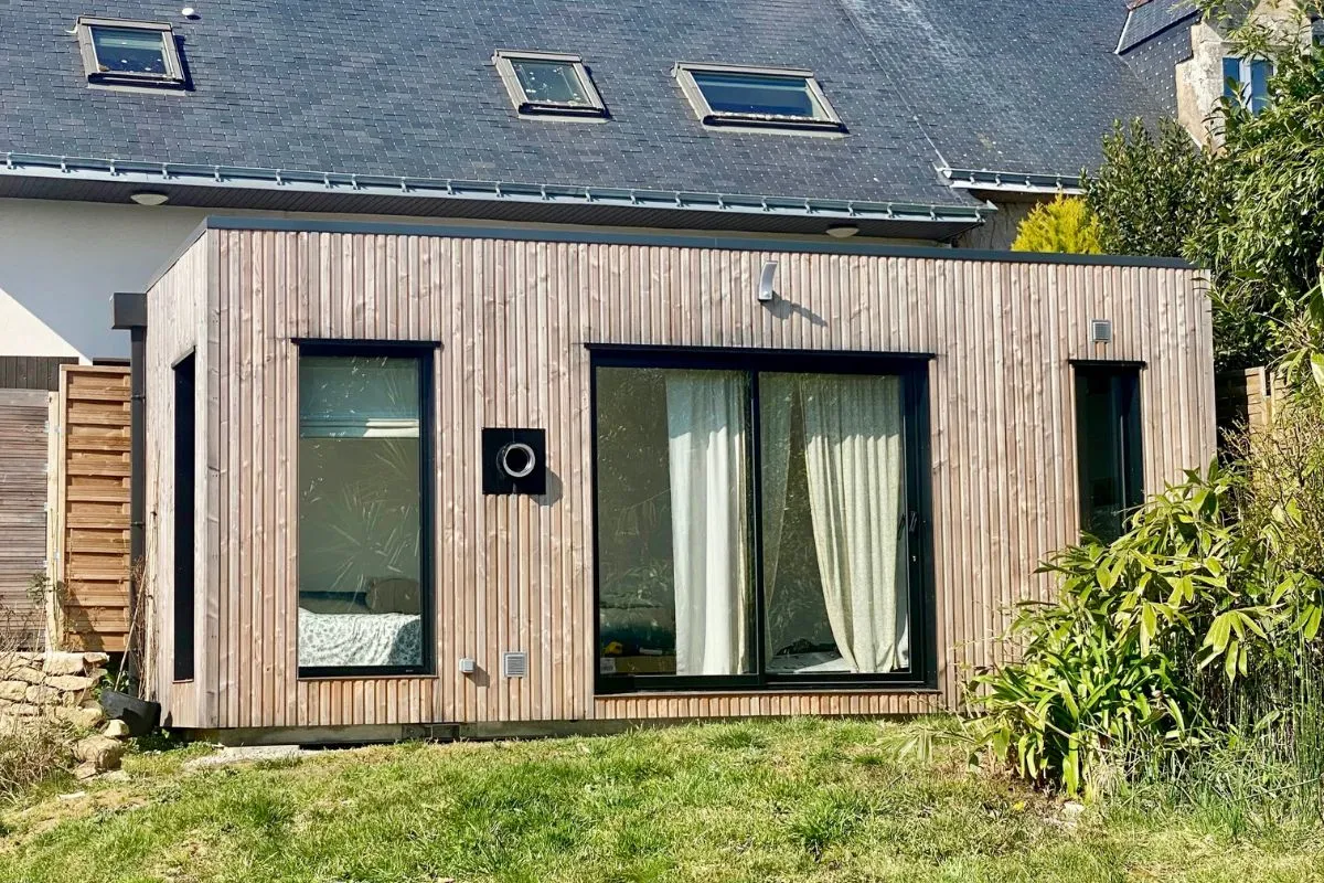 Studio de jardin en bois clair avec grandes fenêtres, intégré à un espace extérieur verdoyant près d'une maison.