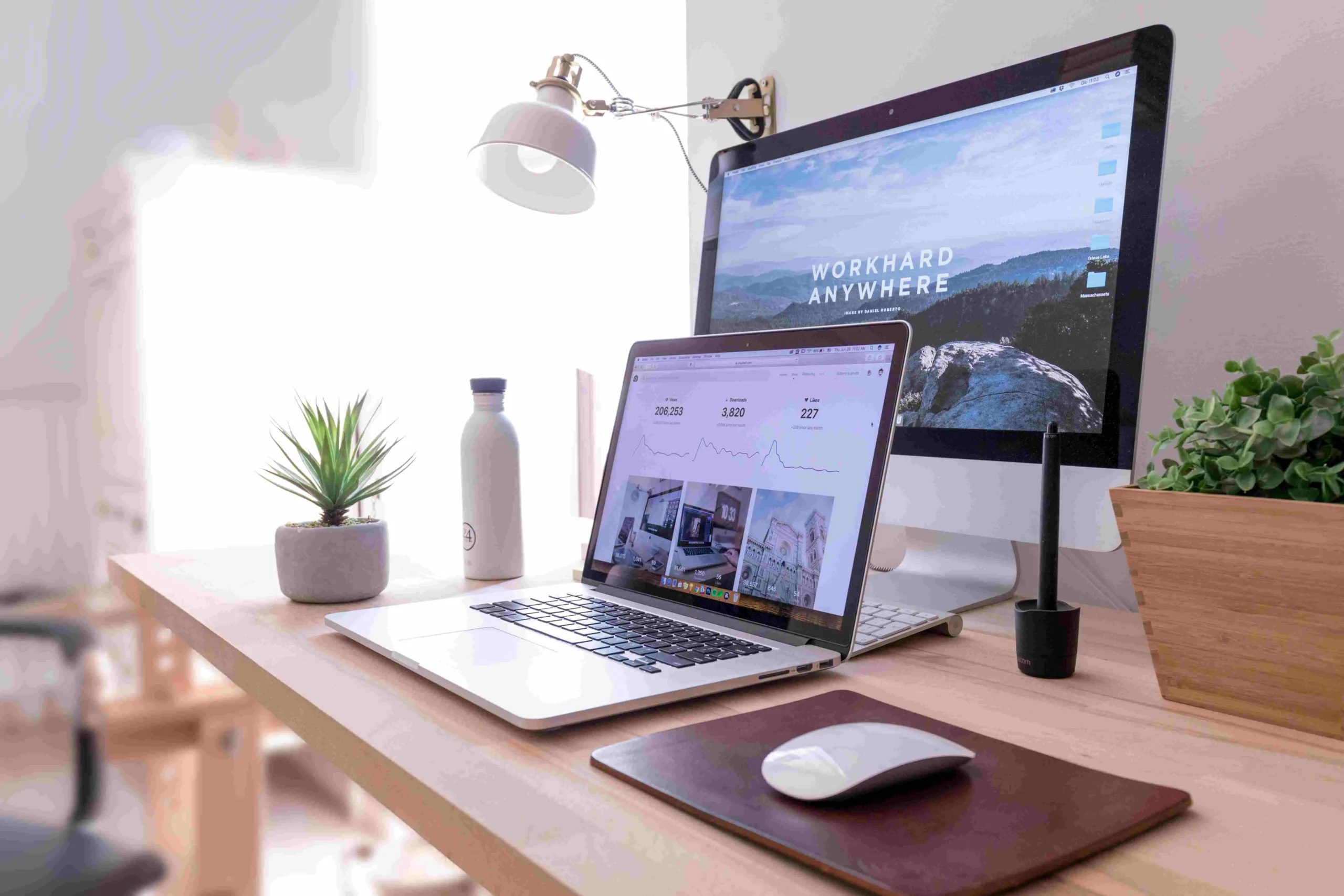 Bureau partagé lumineux et moderne, équipé d'un ordinateur de bureau, d'un ordinateur portable, de plantes vertes et d'accessoires de travail, créant un espace propice à la productivité et au télétravail.
